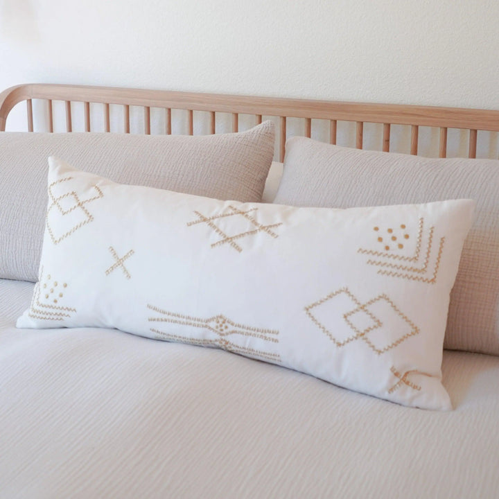 An oversized lumbar pillow with a white cotton cover and gold embroidery design, placed on a minimalist bed. The pillow adds a touch of metallic glamor to the room.
